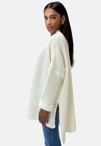 Maglione lavorato a maglia spesso di colore crema con spacchi laterali, maniche lunghe e polsini a coste, abbinato a jeans di denim blu. Design in tessuto texture e trama aperta.