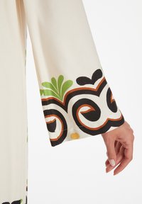 Langsleeve kledingstuk in crèmekleurige stof, met gedurfde zwarte, groene en oranje bloemmotieven aan de zoom van de mouwen en decoratieve stiksels.