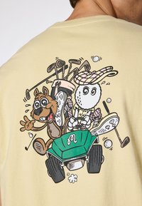 Light yellow t-shirt featuring a graphic of a cartoon character driving a green golf cart, surrounded by golf clubs and a golf ball.