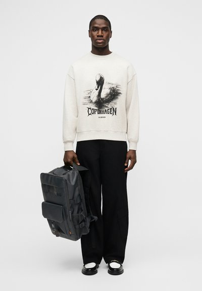 Άνδρας που φοράει λευκό φούτερ με γραφικό κύκνου και το κείμενο "Κοπεγχάγη", μαύρο παντελόνι, μαύρα και λευκά παπούτσια, κρατώντας μεγάλο μαύρο σακίδιο στο πλάι.