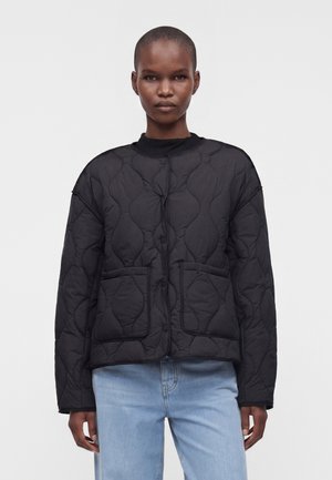 Person står med fronten fremad, iført en sort quiltet jakke med forlommer og lyseblå jeans mod en ensfarvet lys baggrund.