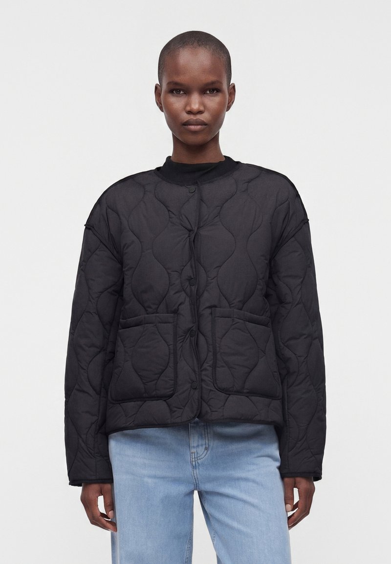 Personne debout face à l'avant, portant une veste noire matelassée avec des poches devant et un jean bleu clair sur un fond uni et clair.