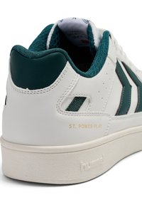 Białe sneakersy z ciemnozielonymi akcentami, z teksturowanym piętą, detalem logo i perforowanym wzorem po boku. Złote litery mówią "ST. POWER PLAY."