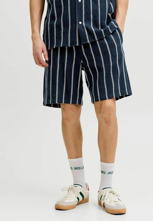 Man draagt marineblauwe korte broek en bijpassend shirt met witte verticale strepen, witte sokken met groene tekst en witte sneakers.