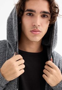 Joven con cabello rizado castaño, vistiendo una sudadera gris sobre una camiseta negra, sosteniendo los bordes de la sudadera cerca de su cara, con expresión neutral.
