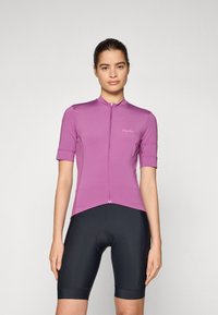 Maillot de cyclisme rose à manches courtes avec une fermeture éclair sur le devant, ourlet arrondi, et logo Rapha. Associé à un cuissard de vélo noir rembourré. Tissu lisse et ajusté.