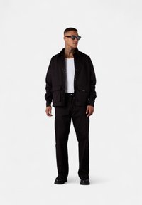 Black lightweight jacket with snap closure, two front pockets, paired with matching black trousers and a white tank top. Black sunglasses.