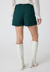Grüne hoch taillierte Shorts mit glatter Textur, die einen sauberen, maßgeschneiderten Schnitt und subtile Falten aufweisen. Weiße kniehohe Stiefel runden das Outfit ab.