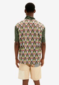 Kortärmad skjorta i grönt tyg med ett repetitivt mönster av röda och blå ovala former, med en kontrasterande texturerad svart ärm.
