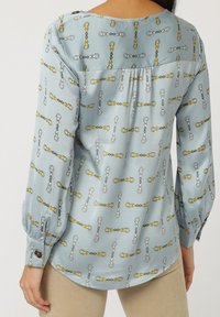 Blusa azul claro con un patrón de cadenas en amarillo y gris. Presenta mangas largas y detalles de botones en los puños. Textura suave.
