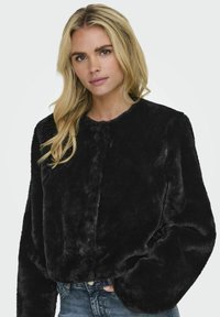 Giacca in pelliccia sintetica nera con scollatura arrotondata e maniche lunghe, caratterizzata da una morbida trama shaggy e un design cropped, indossata sopra a dei jeans.