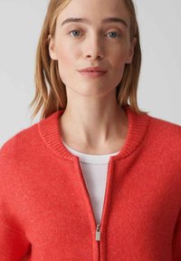 Rode ritsshirt cardigan met ribbelkraag, zachte textuur, iets oversized pasvorm, gedragen over een wit binnenhemd.