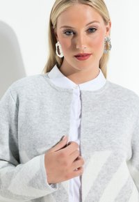 Lichtgrijze cardigan van zacht materiaal, met een ronde halslijn en een patroon in het wit. Lange mouwen en knoopsluiting.