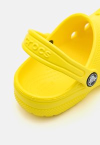 Žute Crocs cipele s podignutom trakom, izrađene od laganog materijala. Imaju ventilacijske otvore i teksturirani potplat za prianjanje. Logo je s bočne strane.