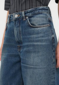 Gros plan sur un jean en denim bleu moyen avec des zones délavées, un bouton frontal et des coutures orange, porté avec une chemise noire à fines rayures.