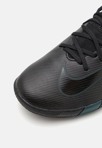 Zapatilla deportiva negra con un upper texturizado, que presenta un logo prominente en un tono oscuro y un acento translucido en color teal cerca de la suela.