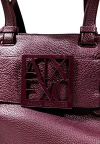 Bolso de mano de cuero burdeos oscuro con acabado texturizado, que presenta una placa rectangular con el logotipo que dice "Armani Exchange". Doble asa.
