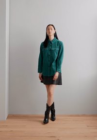 Türkise Cord-Hemden mit Knopfleiste und zwei Brusttaschen, kombiniert mit einem schwarzen Lederrock und schwarzen Cowboy-Stiefeln, vor einer hellen Wand.
