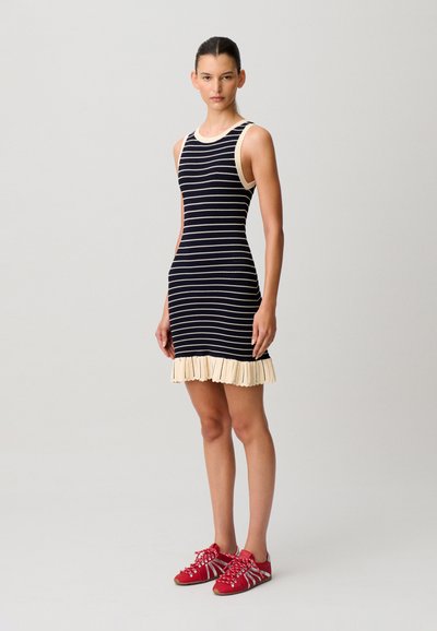 Frau steht, trägt ein ärmelloses schwarz-weiß gestreiftes Kleid mit einem plissierten cremefarbenen Saum und roten Sneakers mit weißen Streifen.