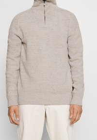 Pull en beige clair avec un col zippé à quart, motif en tricot texturé, poignets et ourlet côtelés. Idéal pour le superposition par temps plus frais.