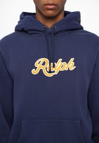 Sudadera azul marino hecha de tela suave, con un bolsillo frontal y un logo bordado de "Ralph" en amarillo en el pecho. Capucha ajustable con cordón.