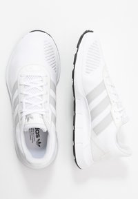 Witte sportschoenen met een bovenwerk van mesh, voorzien van drie grijze strepen en een witte vetersluiting. Getextureerde zool met zwarte accenten.