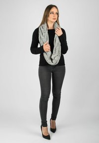 Écharpe en tricot gris en forme d'infini avec un motif de câble texturé, portée lâchement autour du cou sur un haut noir à manches longues et un jean sombre.
