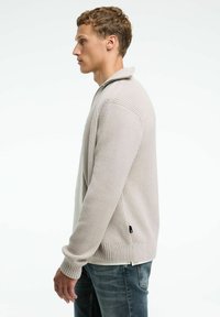 Bežs ribots adījuma džemperis ar augstu apkakli, ar rāvējslēdzēju un sānu izgriezumiem, apvienots ar zilām džinsu biksēm. Sānu skats.