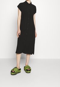 Black button-up dress with short sleeves and side slit, paired with green and black striped fur slide sandals.