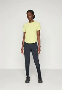 Geel sportt-shirt en zwarte leggings, gecombineerd met zwarte sportschoenen. Het shirt heeft een klein logo aan de voorkant en is gemaakt van lichtgewicht stof.