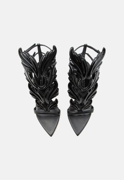 Μαύρα σανδάλια με μυτερή μύτη και ψηλούς ιμάντες στον αστράγαλο, με έντονα, λαμπερά σχέδια φτερωτών φτερών σε λευκό φόντο.