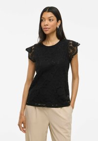 Top in pizzo nero con maniche corte a strascico, caratterizzato da intricati motivi floreali e un collo rotondo. Indossato sopra pantaloni beige.