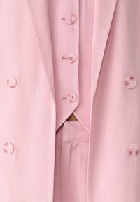 Vaaleanpunainen kaksirivinen bleiseri, jossa on samaa vaaleanpunaista liiviä ja housuja, joissa on pyöreät vaaleanpunaiset napit ja teksturoitu kangas.