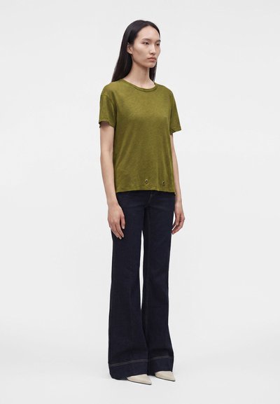 Femme debout, légèrement tournée vers la droite, portant un haut à manches courtes vert olive, un pantalon large foncé et des chaussures blanches pointues sur fond uni.