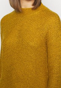 Maglione giallo senape lavorato a maglia con un motivo testurizzato, colletto girocollo a coste e vestibilità leggermente ampia, che mette in evidenza la varia spessore del filato.