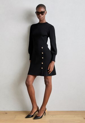 Black dress with ribbed long sleeves, fitted bodice, and a flared skirt featuring gold-tone buttons. Worn with black pointed heels.