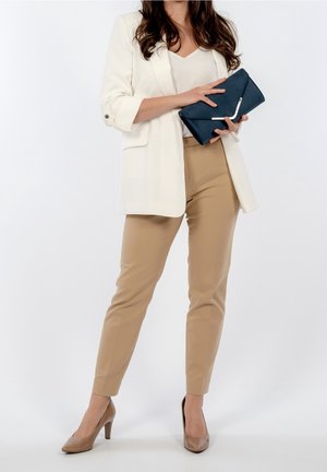 Femme portant un pantalon beige et un blazer blanc, tenant une pochette bleu marine d'une main sur un fond uni.
