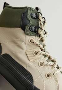 Beige Stoff-Hohe Sneaker mit grünen Akzenten, strukturiertem Gummisohle und schwarzem Zubehör. Merkmale sind geflochtene Schnürsenkel und genähtes Detail.