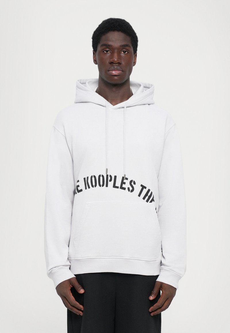 Junger Mann in einem weißen Kapuzenpullover mit schwarzem Text auf der Brust und schwarzen Hosen, der vor einem schlichten hellen Hintergrund steht.