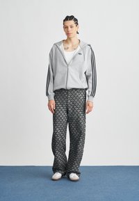 Άτομο που στέκεται σε μπλε χαλί φορώντας γκρι φούτερ με φερμουάρ και μαύρες ρίγες, καθώς και μαύρο παντελόνι φαρδιά μπατζάκια με σχέδιο, και λευκά αθλητικά παπούτσια.