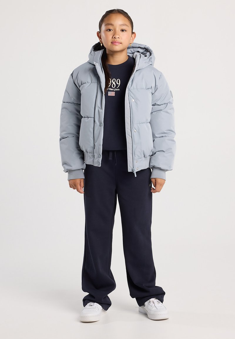 Junge Person trägt eine hellgraue Steppjacke über einem schwarzen Sweatshirt und schwarzen Hosen mit weißen Sneakers, steht vor einem einfarbigen Hintergrund.