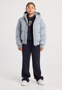 Ein junges Mädchen steht in einer hellblauen Daunenjacke, dunkelblauen Jogginghosen, einem dunkelblauen Sweatshirt mit dem Aufdruck "1989" und weißen Turnschuhen.