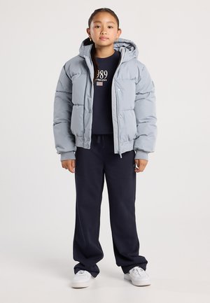 Jeune fille debout portant une veste en doudoune bleu clair, un pantalon de survêtement bleu marine, un sweat-shirt bleu marine avec le texte "1989", et des baskets blanches.