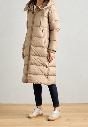 Femme portant un manteau matelassé beige mi-long avec capuche, jean slim foncé et baskets rose pâle et argent, debout sur un sol en bois.