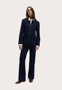 Traje a rayas azul marino hecho de tela estructurada; chaqueta entallada con cremallera, combinada con pantalones acampanados. Se destacan los acentos de cuello blanco.