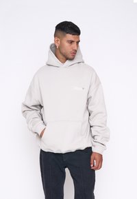 Sweat à capuche gris avec une poche kangourou, des bords côtelés et un cordon de serrage pour la capuche. Présente une broderie de logo discrète sur la poitrine. Texture de tissu douce.