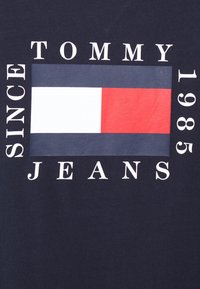 Navy blue T-shirt featuring a large logo with red, white, and grey stripes, and text reading "TOMMY JEANS SINCE 1985" in white lettering.