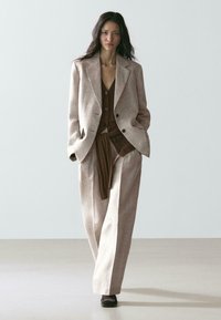 Kvinde står iført beige oversized dragt med vidde bukser og en brun knappet cardigan under, hænderne i lommerne.