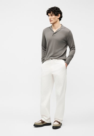 Homme aux cheveux bouclés portant une chemise grise à manches longues, un pantalon blanc et des chaussures beige à lacets, debout les mains dans les poches.