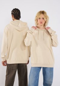 Beige hooded sweatshirt with a front pocket, loose fit, long sleeves, and drawstring. Worn by two models from front and back.